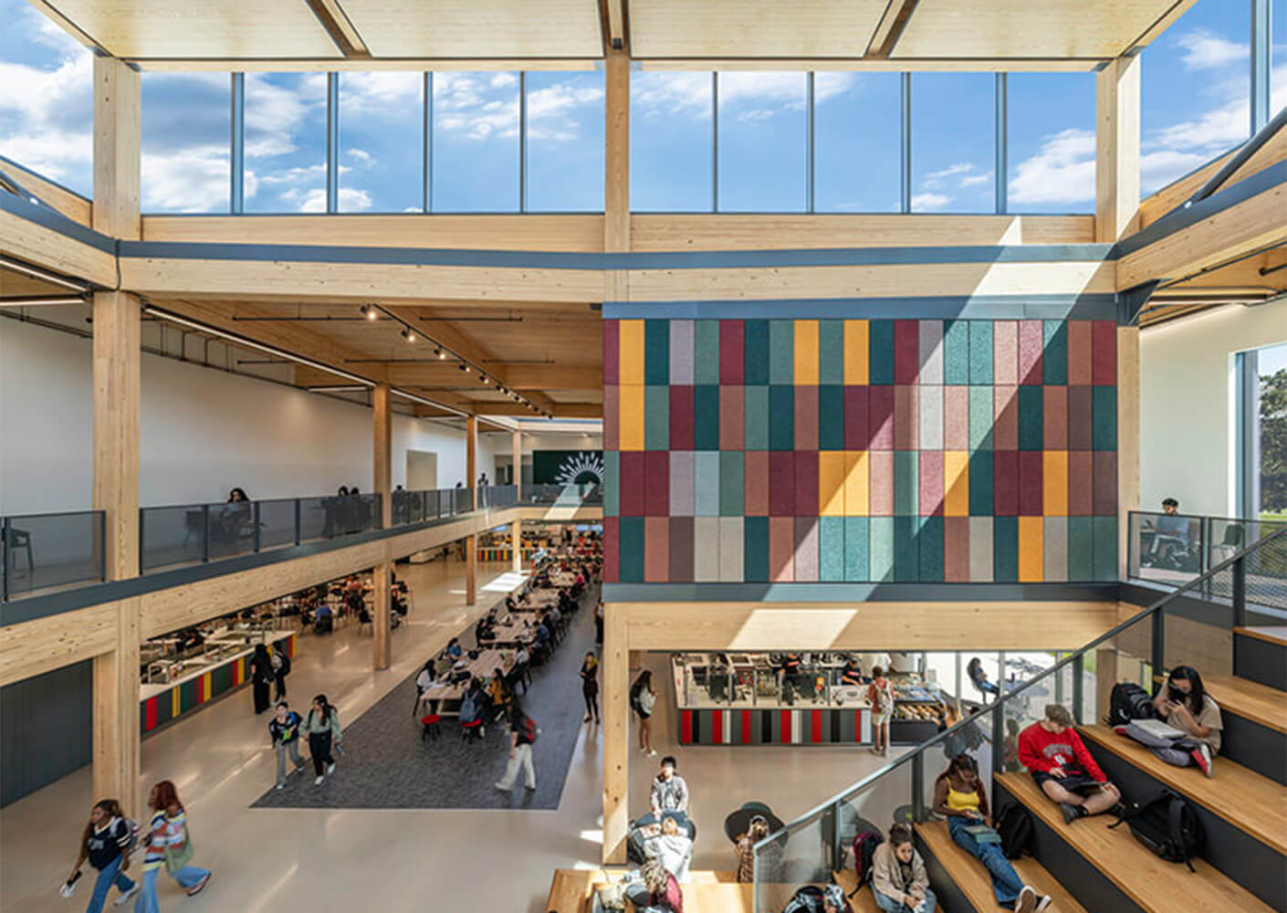A spacious interior with wooden beams, colorful wall art, and students interacting in a dining area and study spaces.