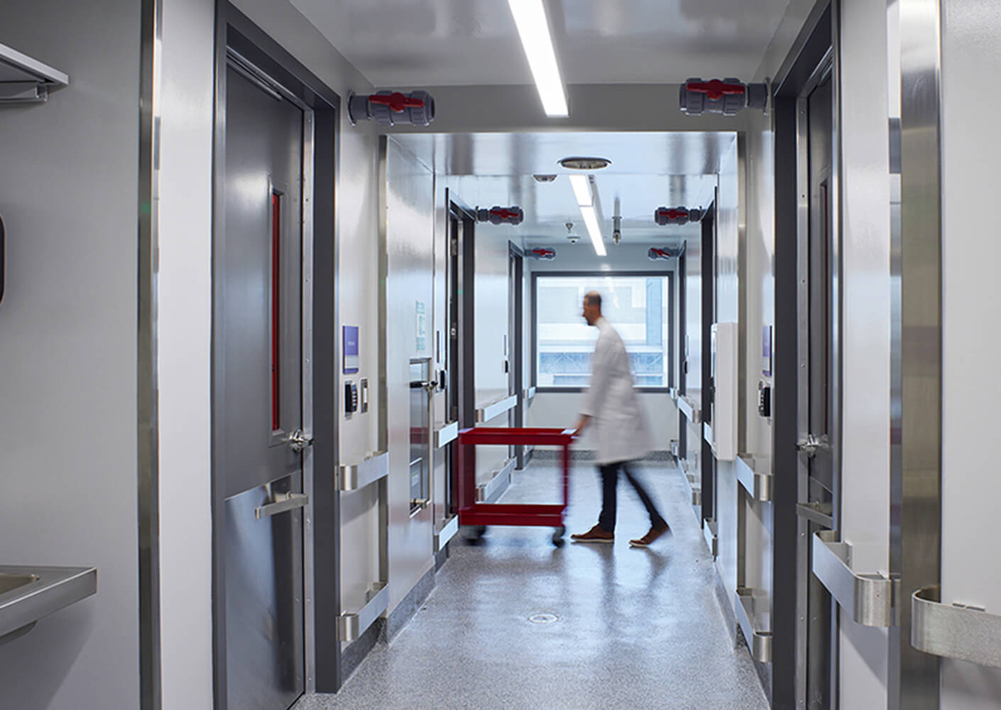 A healthcare professional walks down a corridor, pushing a red cart past doors and windows in a clinical setting.