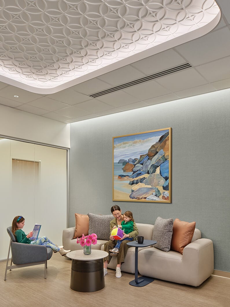 A welcoming waiting area featuring a textured ceiling, a sofa, a coffee table with flowers, and a mother with a child. A girl reads nearby.