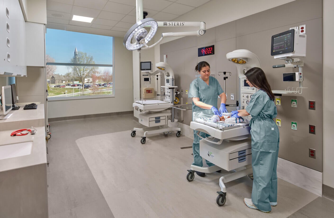 Two healthcare professionals in scrubs tending to a newborn in a neonatal care unit, equipped with medical technology and natural light from a window.
