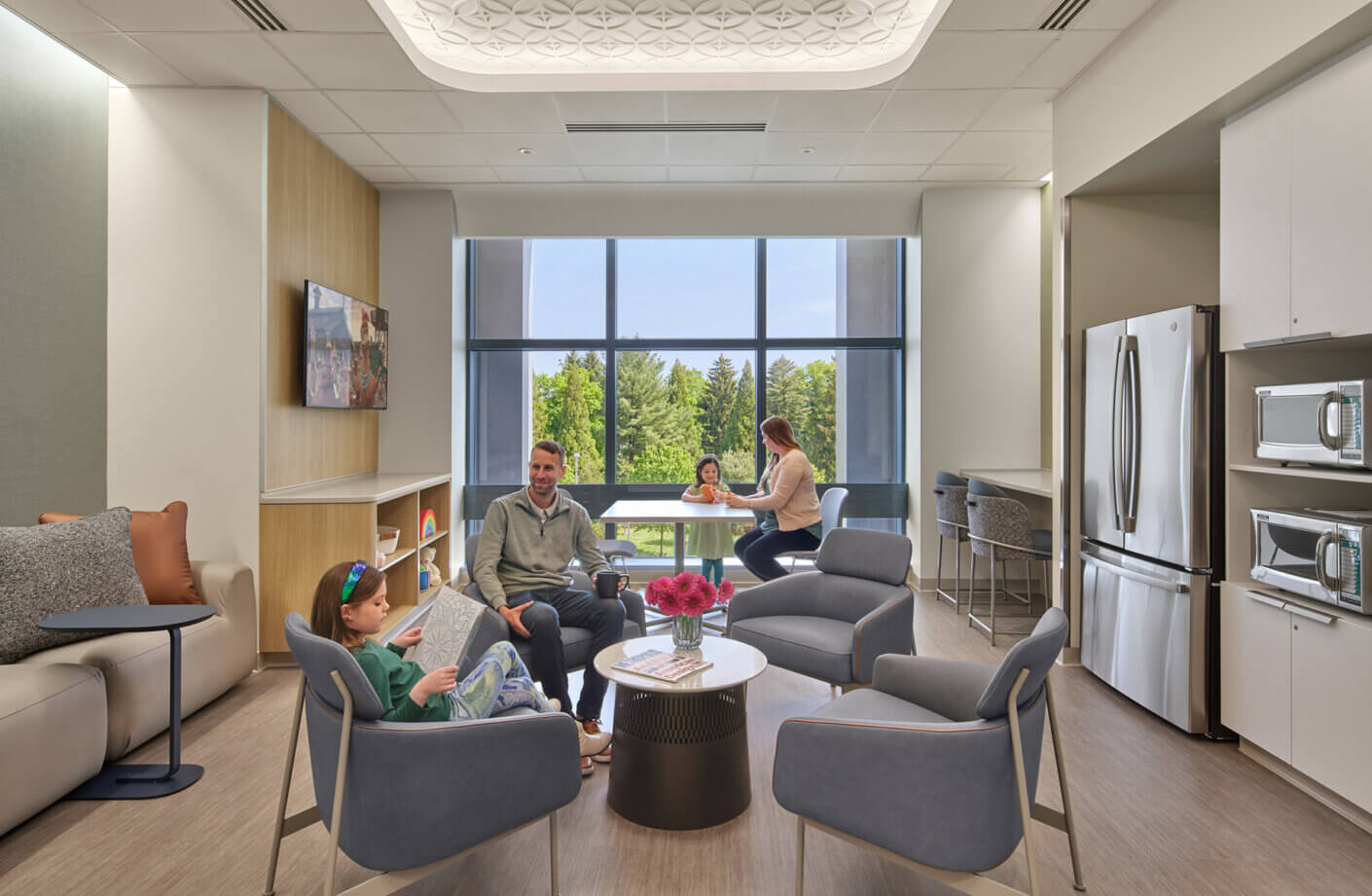 Family lounge area with comfortable seating, a viewing window, and a kitchenette. Children engaged in activities; adults relax and converse.