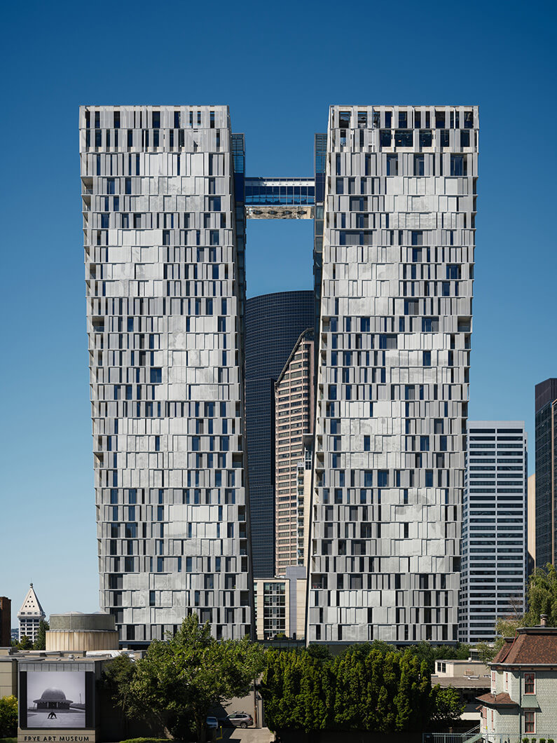 Two tall buildings featuring unique facades and a bridge connecting them, set against a clear blue sky in an urban environment.