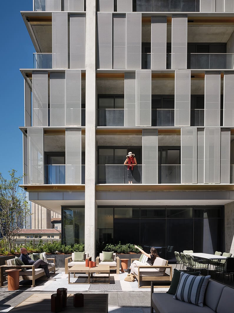 Two people relax in a courtyard with lounge furniture, while a woman leans over a balcony. The building features horizontal metal slats and glass.