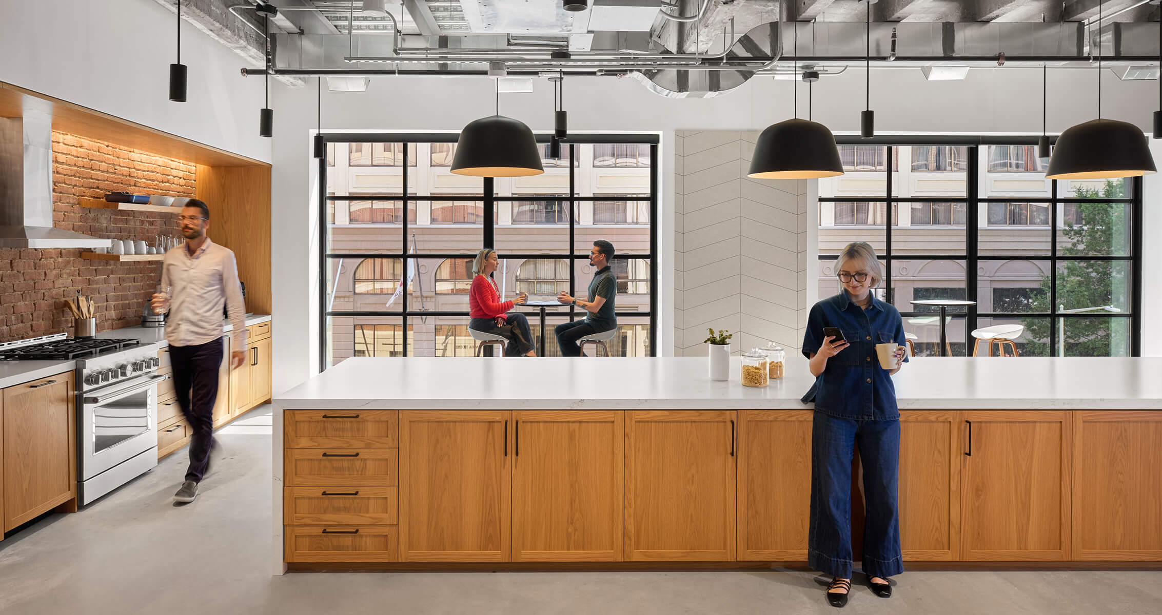 Bright kitchen space with wood cabinets and large windows. A man walks by, while two people sit at a table discussing. A woman checks her phone.