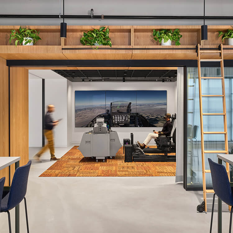 An office space featuring a flight simulator, with a person seated and monitors displaying a flight scene, surrounded by plants and wooden accents.