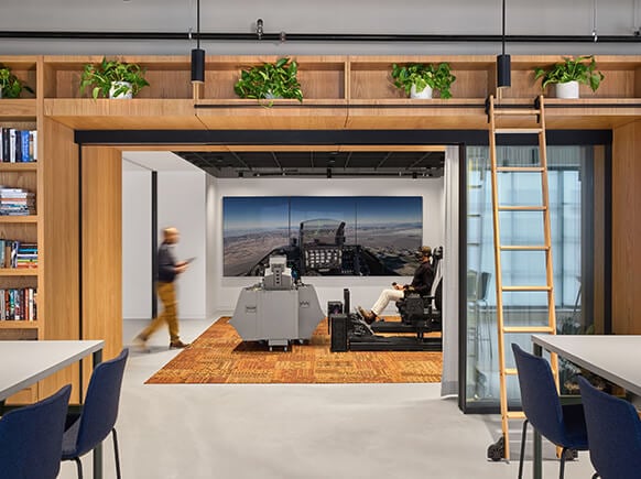 Interior office space featuring seating areas, large screens, and greenery. A person walks by while another is seated at a control station.