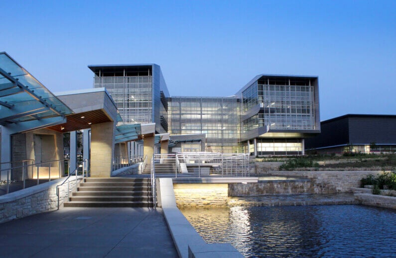 Architectural building with multiple levels, large glass windows, and landscaped surroundings near water. Captured at dusk, showcasing lighting.