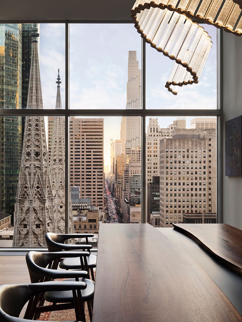View from a high-rise interior featuring cityscape with skyscrapers, spires, and a wooden table with chairs, illuminated by evening light.