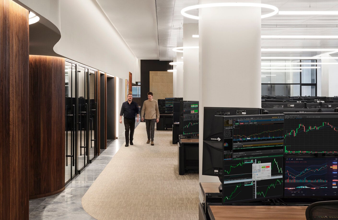Two professionals walk through a bright office space featuring multiple computer desks with financial market displays and a modern interior design.
