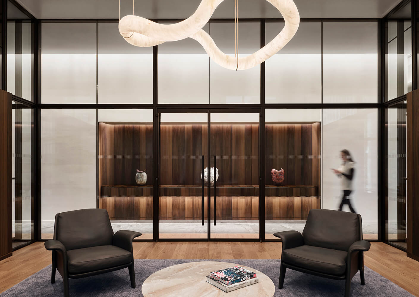 Contemporary lobby featuring glass walls, stylish furniture, and decorative art pieces, with a person walking in the background.