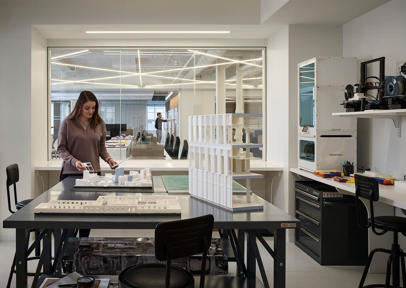 Female architect examining building models at a workspace, with design tools and a 3D printer visible in a well-lit office environment.