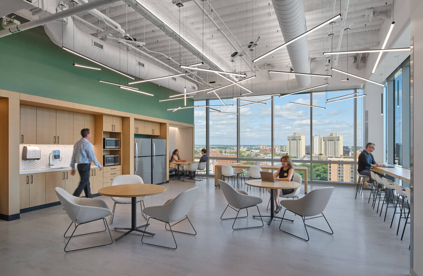 Bright office break room featuring seating areas, kitchen amenities, and large windows with city views. People engaged in work and conversation.