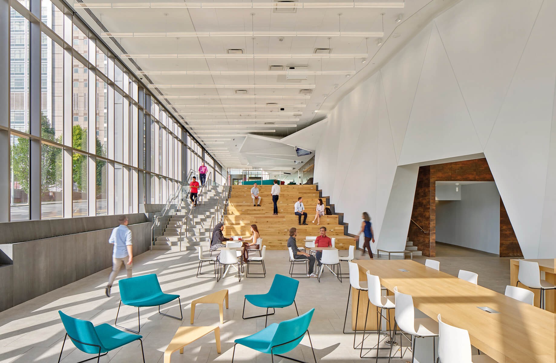 Spacious interior space with large windows, seating areas, and people interacting in a collaborative environment. Stairs lead to an upper level.