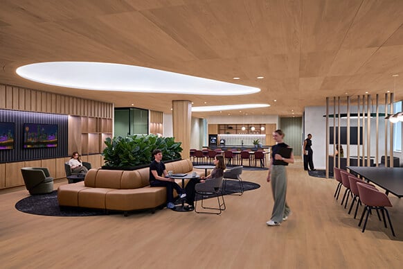 Office lounge area featuring wooden elements, seating arrangements, lush greenery, and a kitchenette with people engaged in various activities.