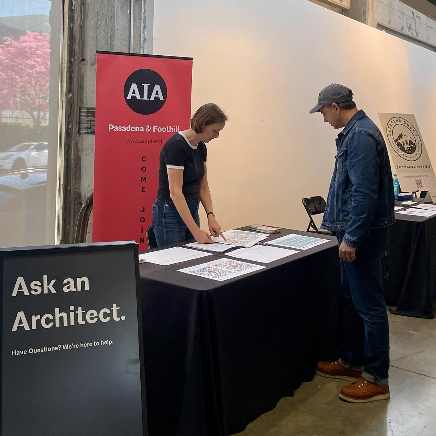 AIA booth at an event with a woman discussing plans with a visitor. Sign reads Ask an Architect inviting questions and interaction.